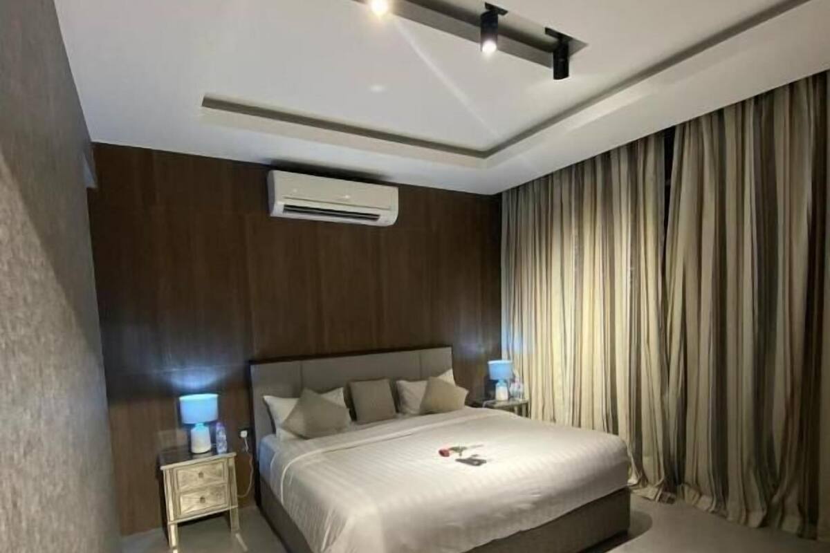 A modern bedroom with a large bed, bedside lamps, an air conditioning unit, and a ceiling light fixture.