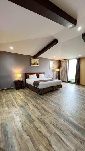 A bedroom with a large bed, wooden flooring, a sloped ceiling, and a window with curtains.