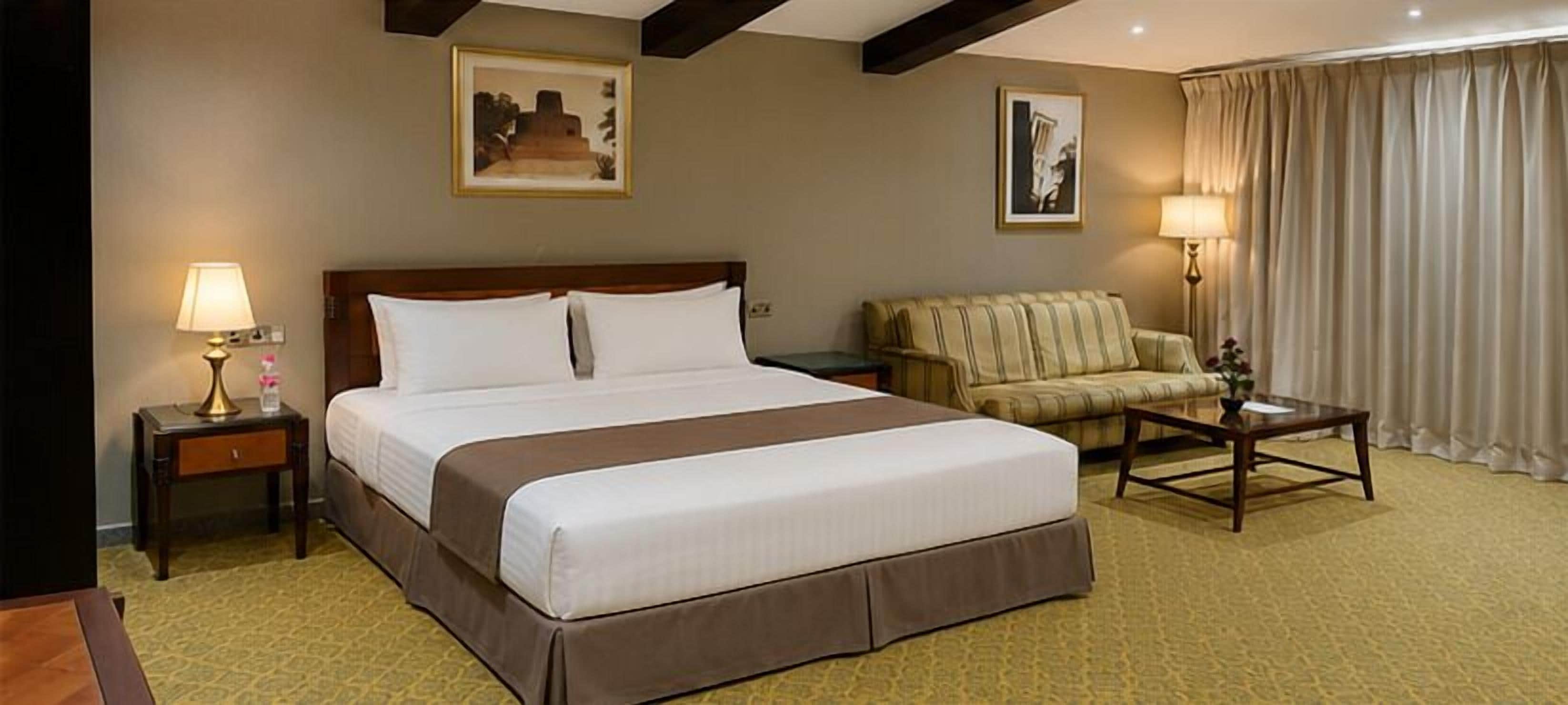 Quarto de hotel com uma cama grande, um sofá, uma mesa de centro e duas luminárias.