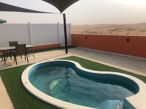 A swimming pool with a unique shape, surrounded by artificial grass, a white fence, and a view of a desert landscape.