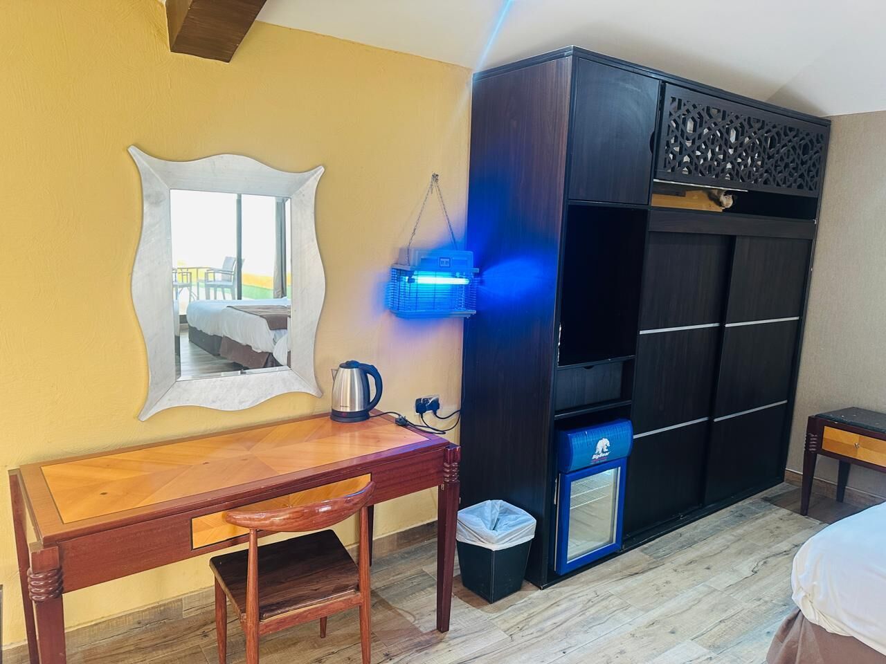 A room with a wooden desk, a chair, a mirror, a wardrobe, a small trash bin, and a heater.