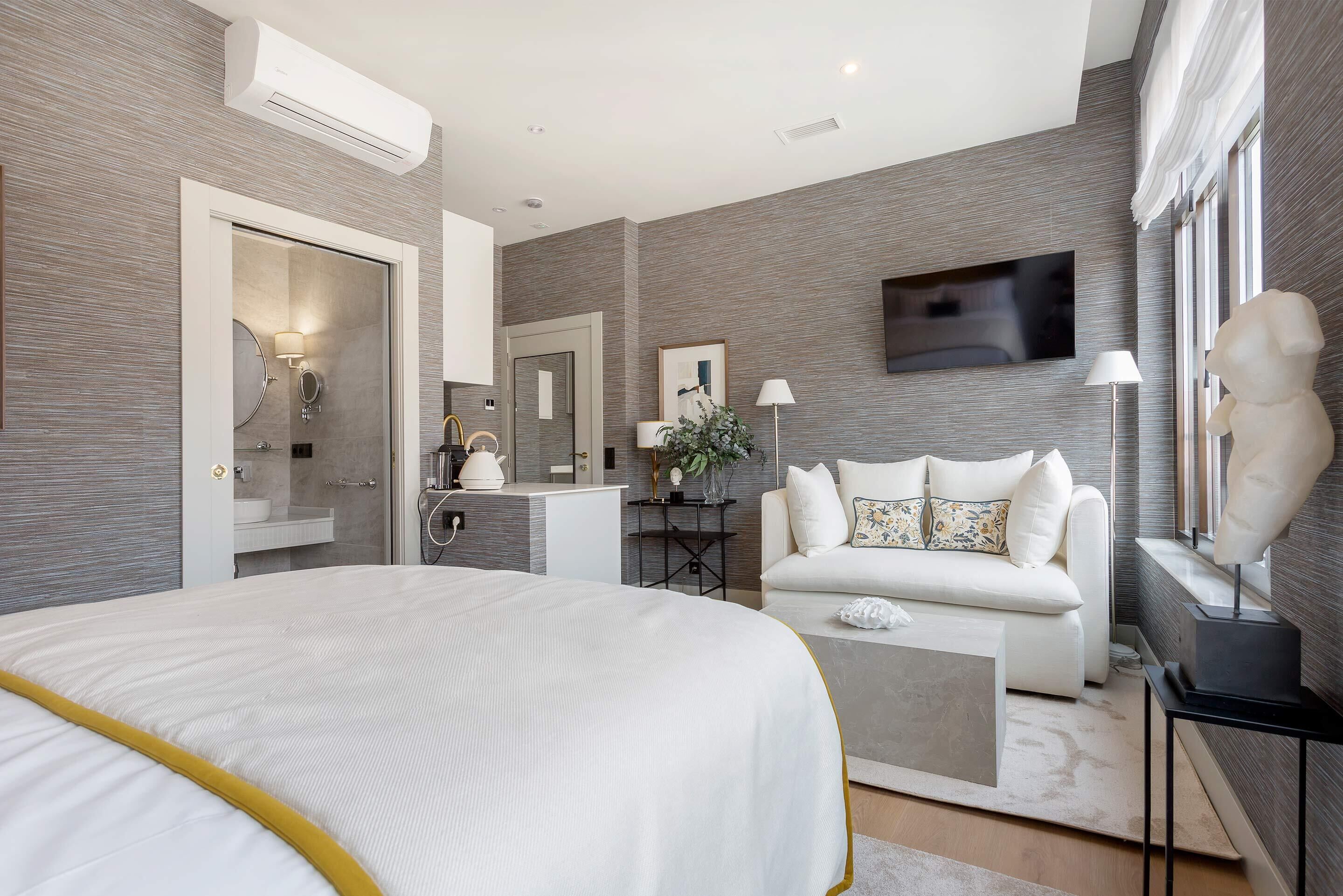 A modern bedroom with a large bed, a sofa, a TV, and a bathroom with a mirror and sink.