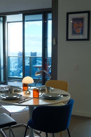 A dining area with a table set for two, a lamp, and a view of a cityscape through large windows. - Elevated Bauhaus 2BR with Bay Views (Melbourne)