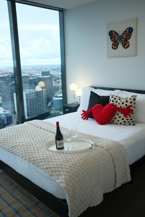 A hotel room with a large window, a bed with a white blanket, a red heart-shaped pillow, a butterfly painting on the wall, and a bottle of wine on the bedside table. - Elevated Bauhaus 2BR with Bay Views (Melbourne)