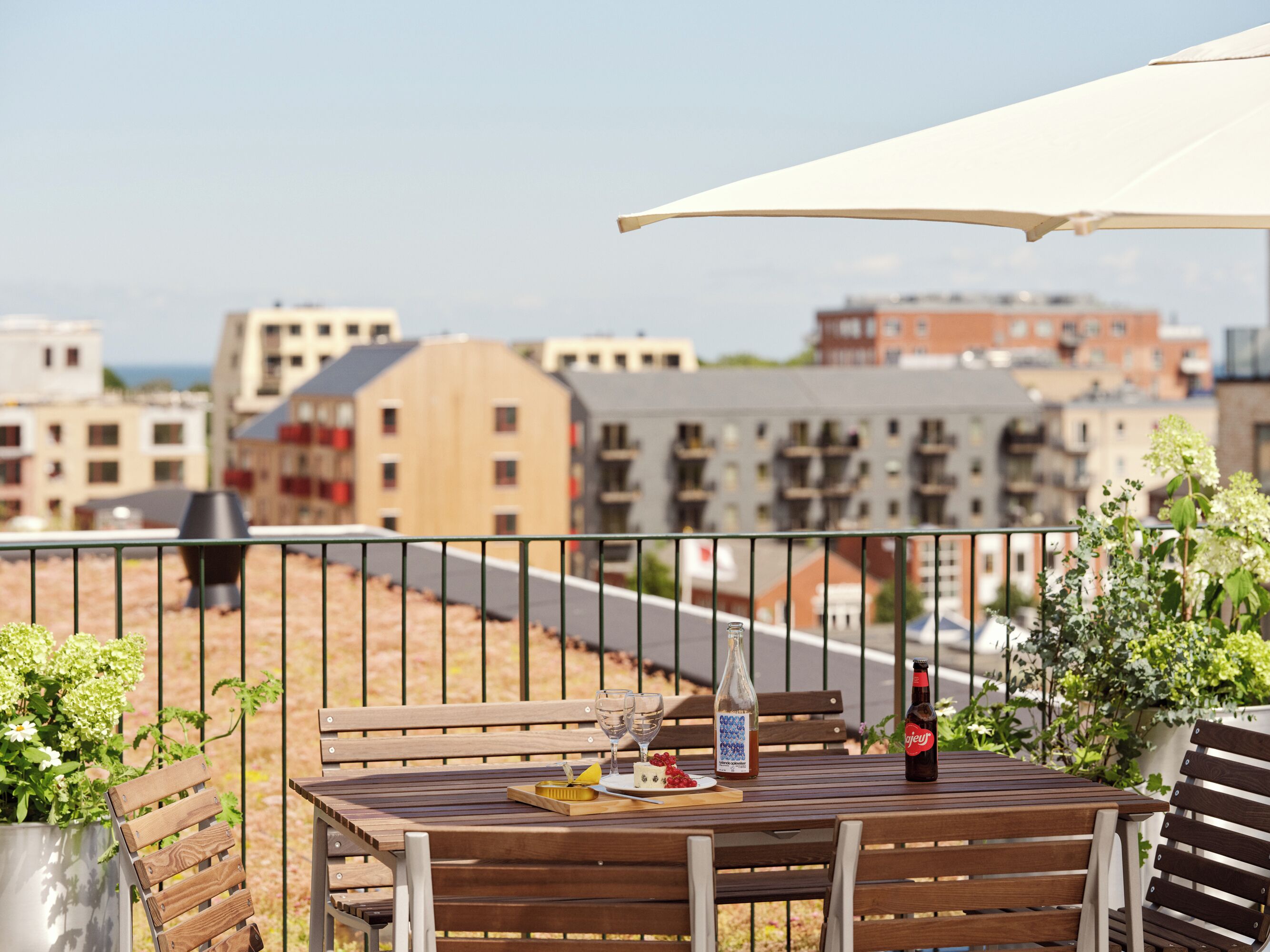 A wooden table with a bottle, a glass, and a plate of food on a balcony with a view of buildings.