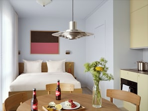A hotel room with a bed, a dining table with a bottle and a plate of food, a vase with flowers, and a kitchen area.