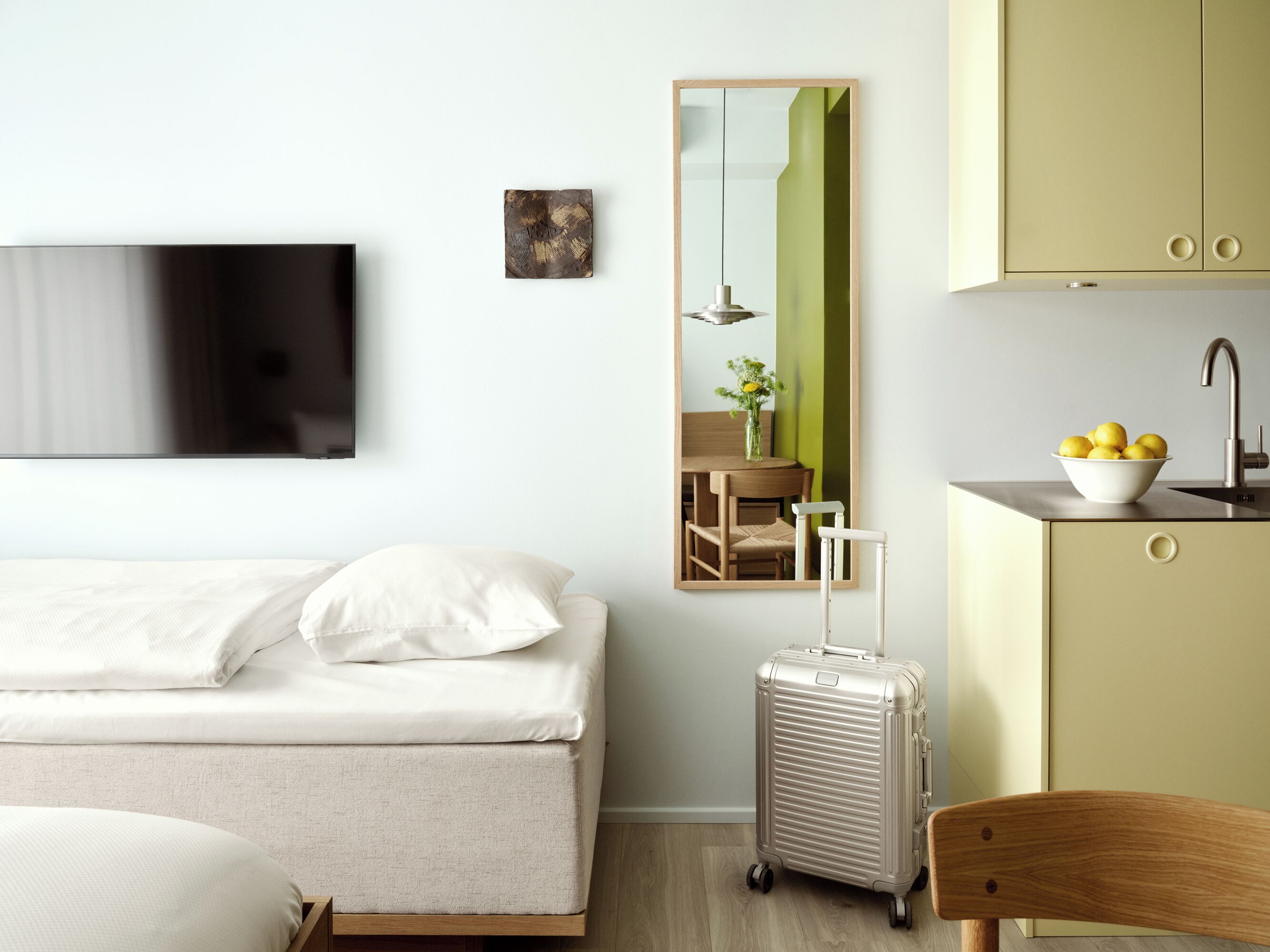 A hotel room with a bed, a mirror, a small kitchenette, and a suitcase.