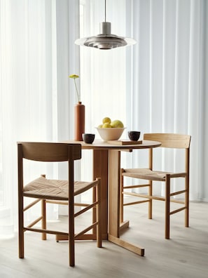 Un coin repas avec une table en bois, deux chaises, un saladier de fruits et un luminaire suspendu.