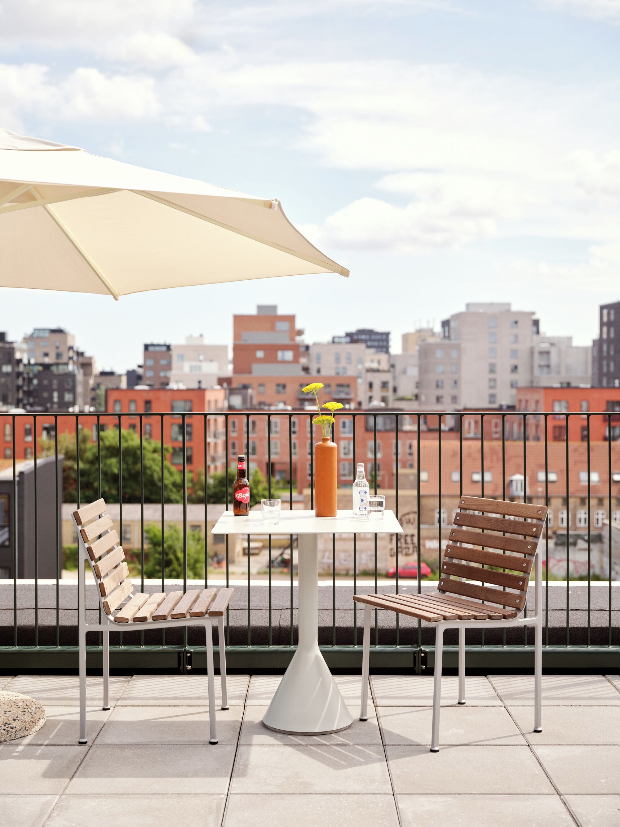 A balcony with two wooden chairs, a table with a bottle and glasses, and a large umbrella.