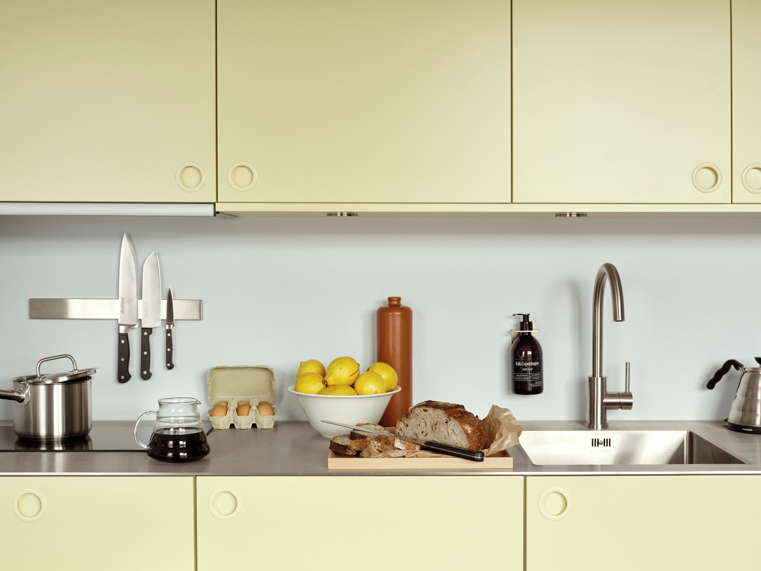 A kitchen with a stainless steel sink, a knife block, a bowl of lemons, eggs, a loaf of bread, and a bottle of oil.