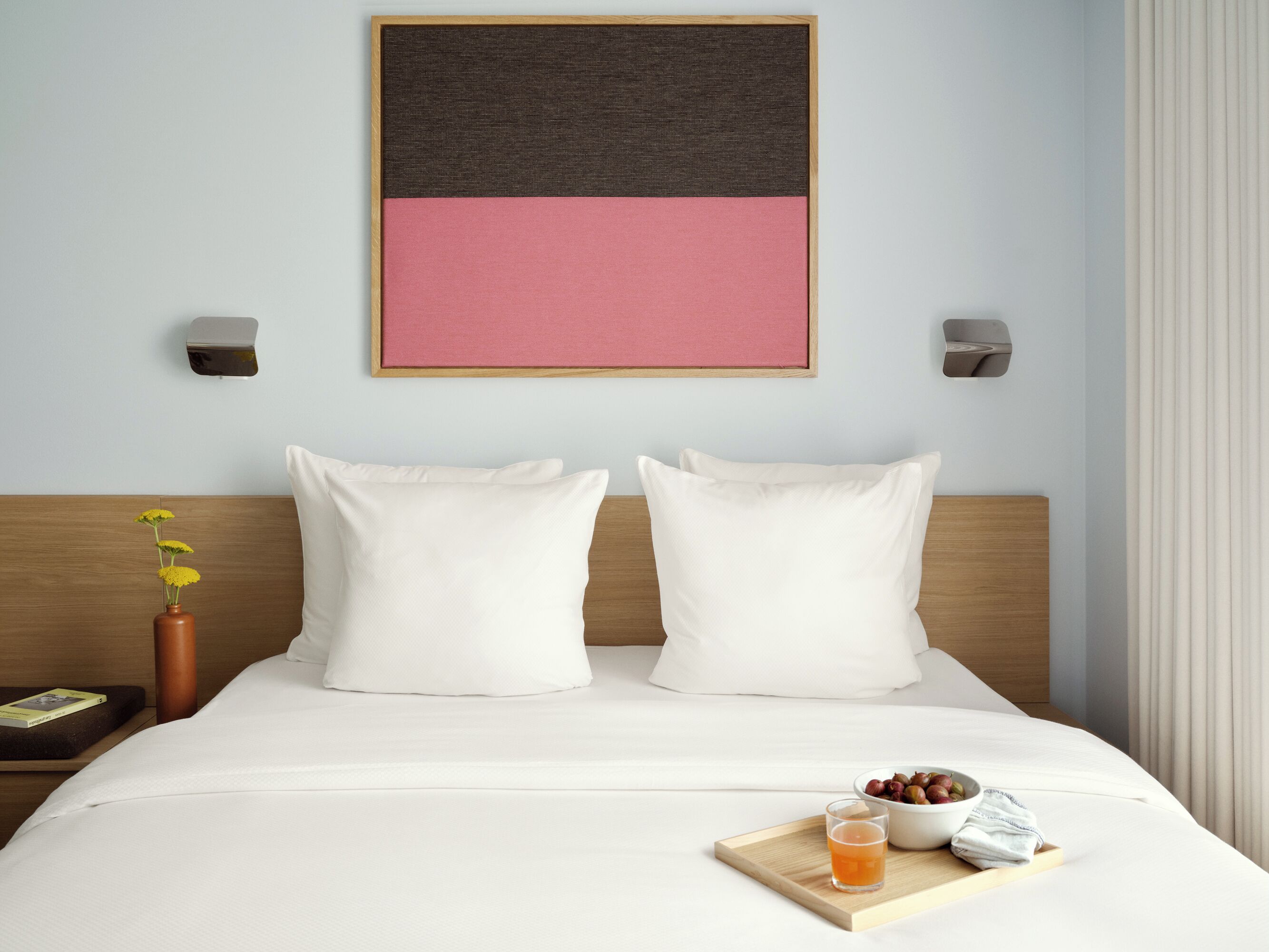 Une chambre d’hôtel avec un lit, des oreillers, un vase de fleurs, une table de chevet avec un livre et un plateau avec une boisson et des en-cas.