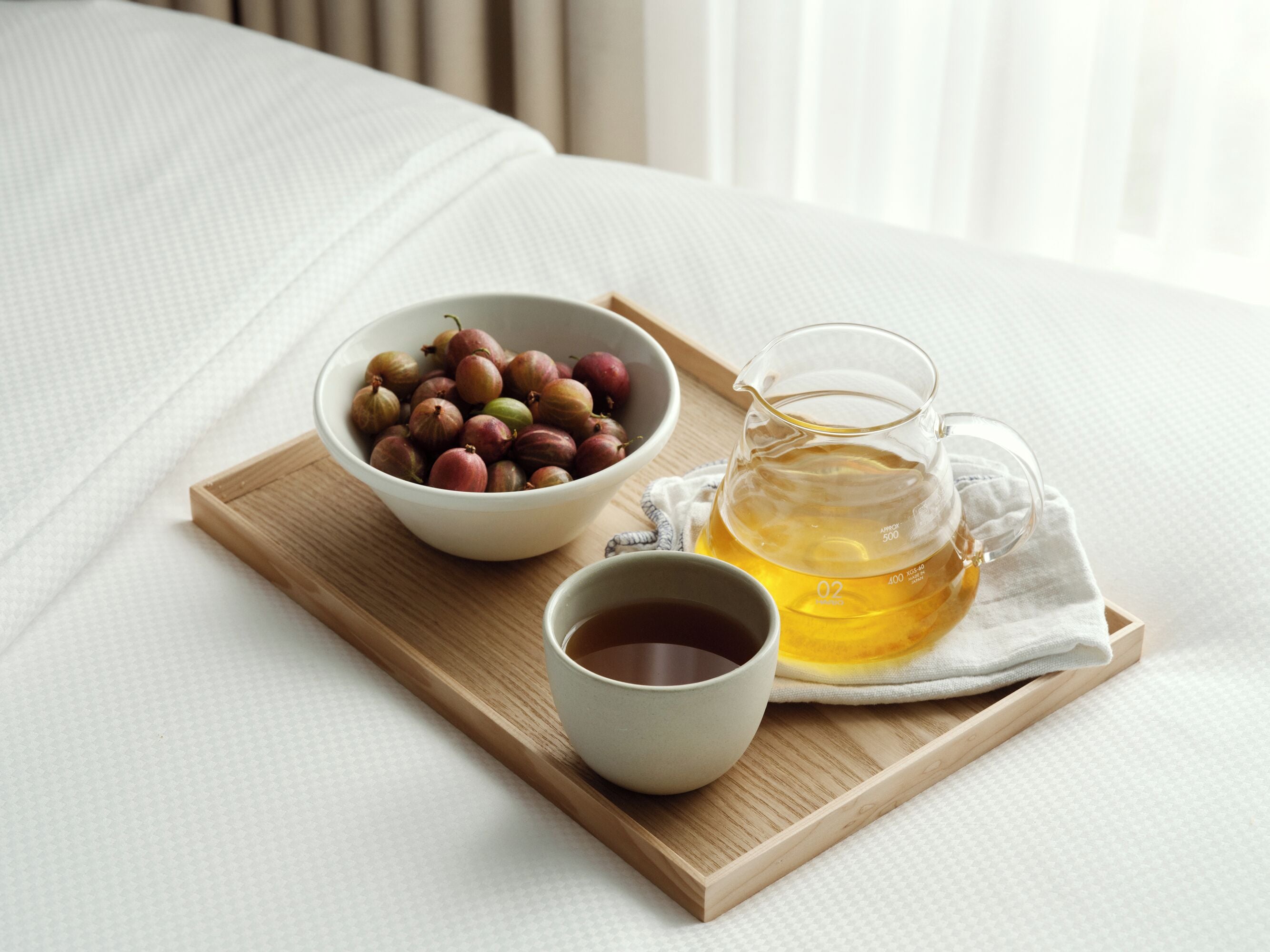A wooden tray with a bowl of dried fruits, a glass pitcher of tea, and a cup of tea on a bed.