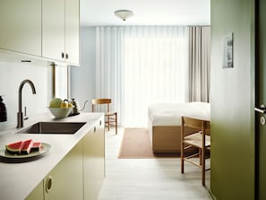 Une chambre d’hôtel de taille réduite, équipée d’une kitchenette, d’un lit, d’une chaise et d’une fenêtre avec des rideaux.