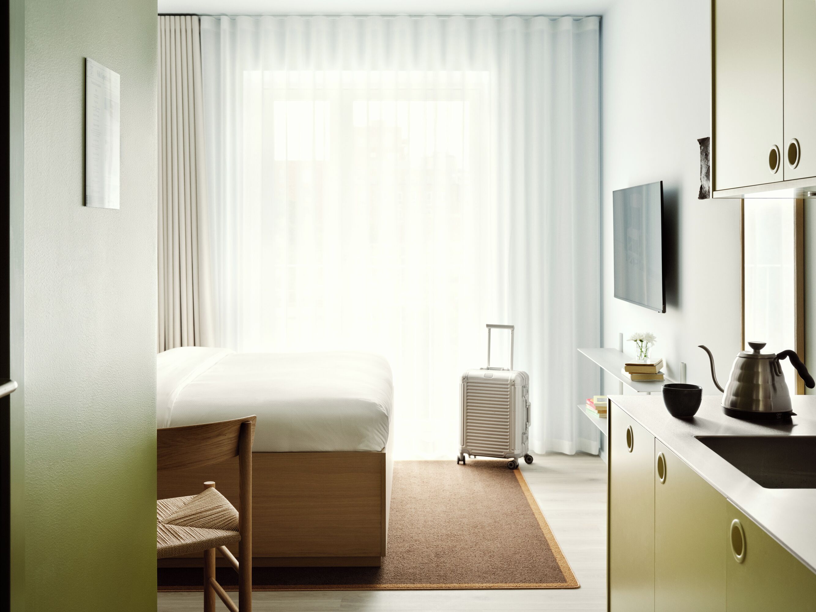 A hotel room with a bed, a chair, a suitcase, a desk with a kettle, and a television.