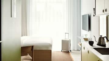 A hotel room with a bed, a chair, a suitcase, a desk with a kettle, and a television.