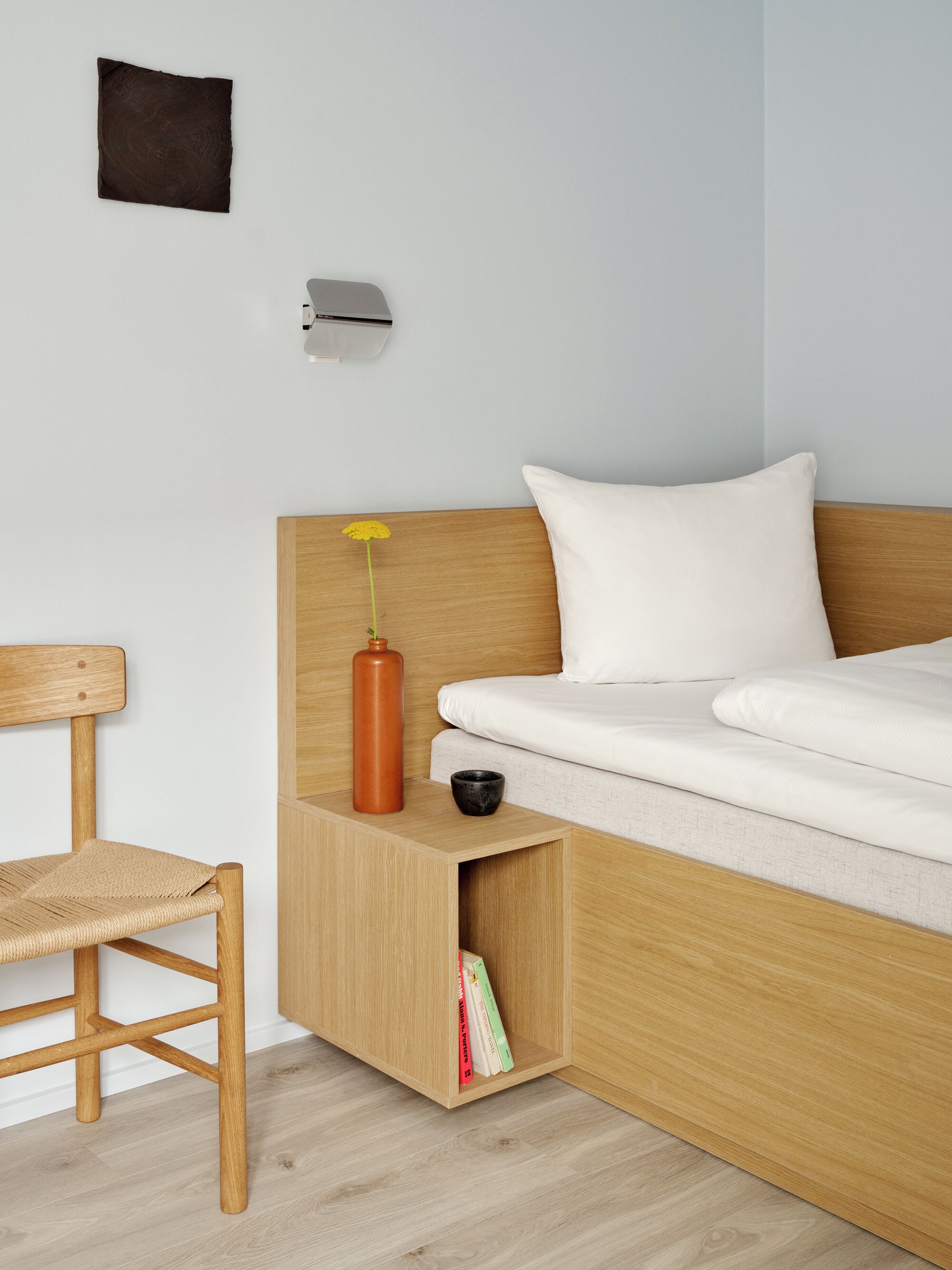 Un dormitorio moderno con cabecera de madera, colchón blanco, una mesita de noche con un florero y libros, y una silla.