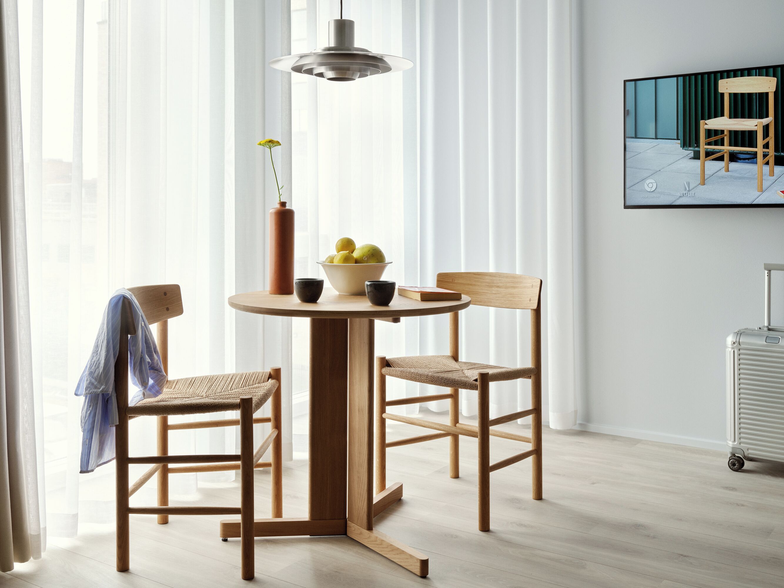 A modern dining area with a round table, two wooden chairs, a vase with flowers, and a bowl of fruit.