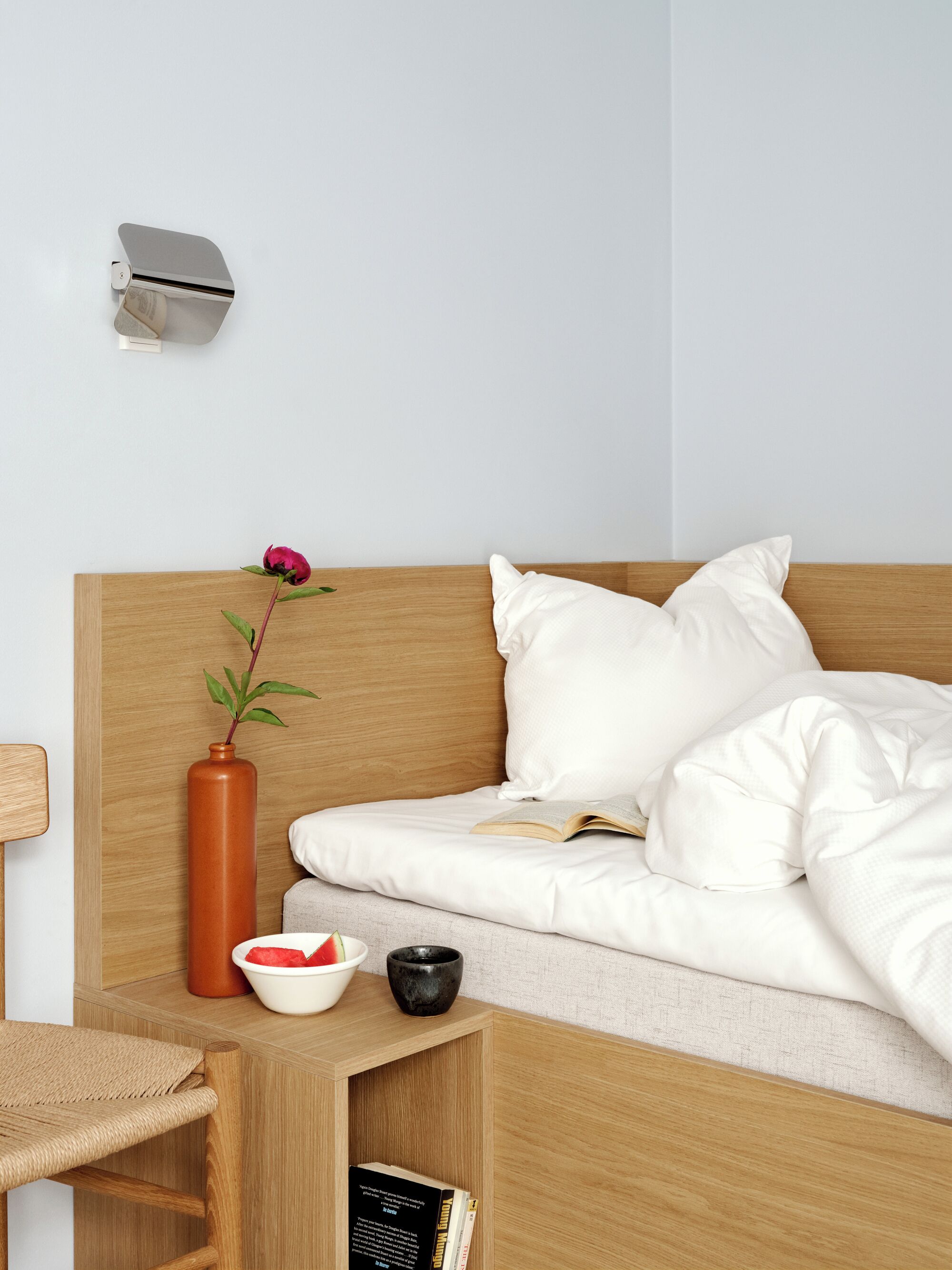 A neatly made bed with white bedding, a wooden headboard, a small bedside table with a vase and a bowl, and a wall-mounted light fixture.
