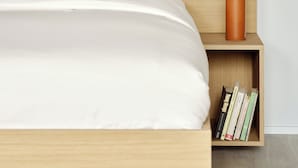 Ein ordentlich bezogenes Bett mit weißer Bettwäsche, ein hölzerner Kopfseitenverkleidung, ein kleiner Nachttisch mit einer Vase weißer Blumen und ein Bücherregal.