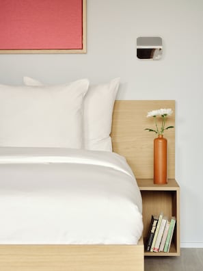 A neatly made bed with white bedding, a wooden headboard, a small wooden nightstand with a vase of white flowers, and a shelf with books.