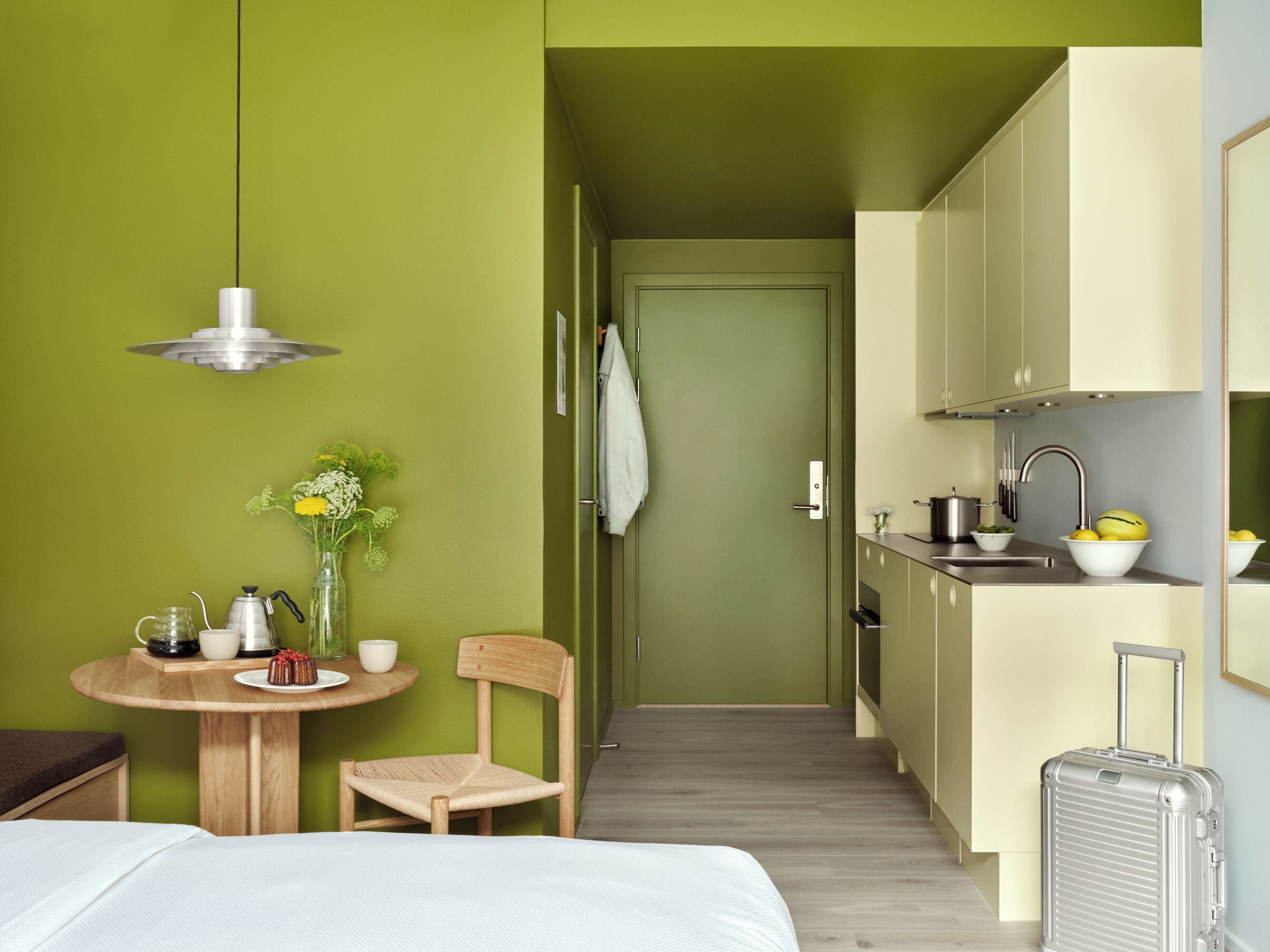 A compact hotel room with a bed, a small round table with a chair, a kitchenette, and a suitcase.