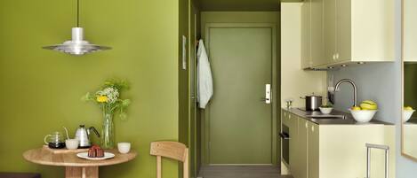 A compact hotel room with a bed, a small round table with a chair, a kitchenette, and a suitcase.