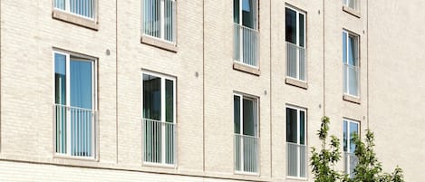 Un edificio de varios pisos con fachada de ladrillo, ventanas grandes con marcos blancos y un patio con plantas y mobiliario exterior.
