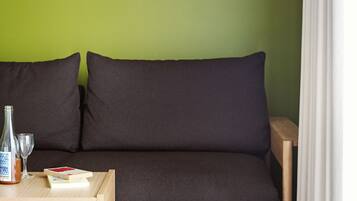 A room with a green wall, a dark sofa, a wooden side table with a bottle and glasses, and a small framed picture.
