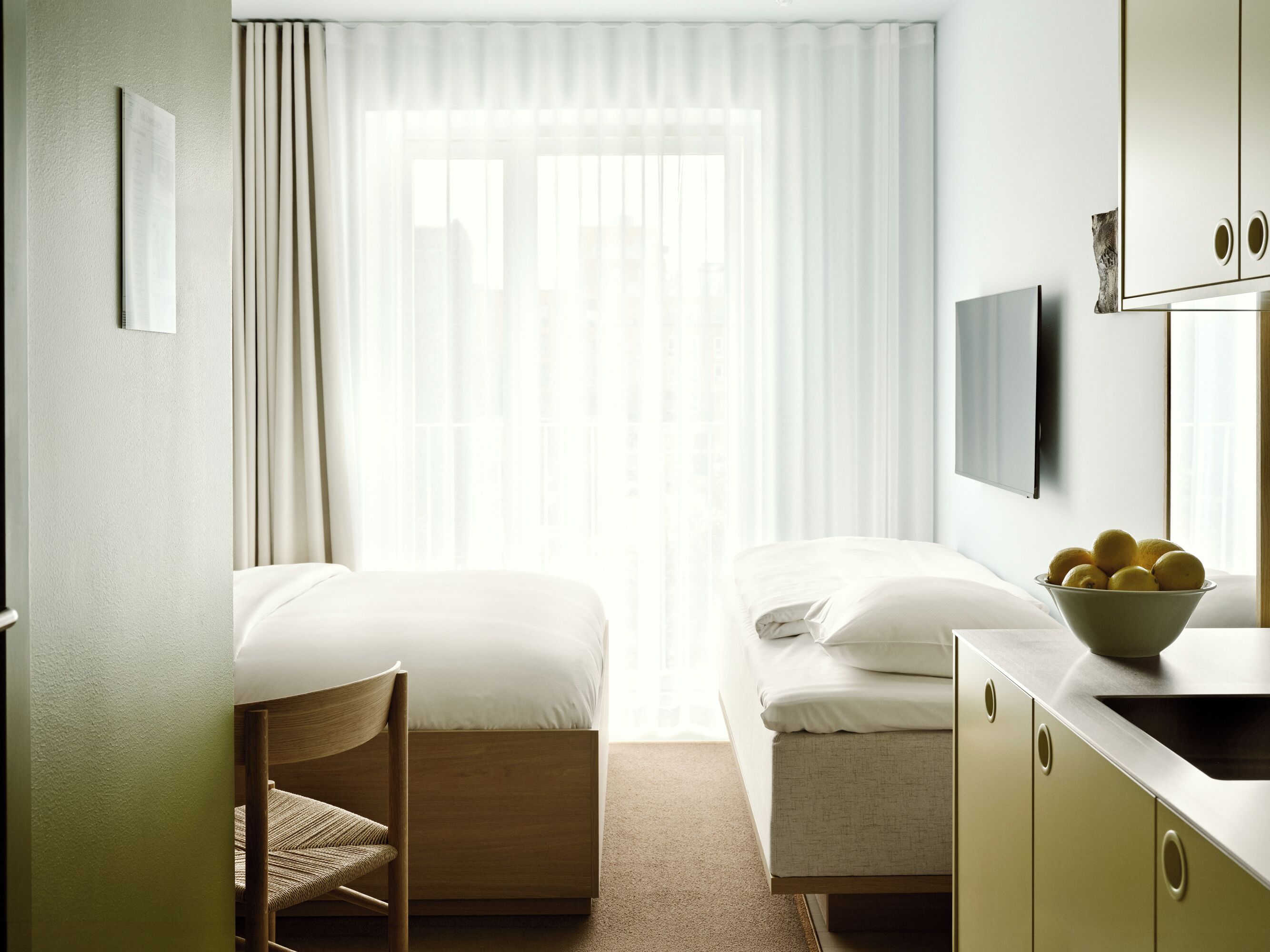 A hotel room with a bed, a chair, a bowl of fruit, a television, and a window with curtains.