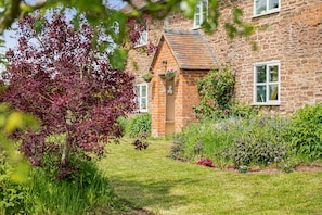 A stone building with a garden in front, a small brick structure with a pitched roof, and a window with a white frame.