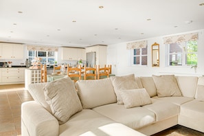 A modern living room with a beige sofa set, a dining area with wooden chairs, and a kitchen with white cabinets.