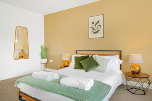 Habitación de hotel con cama, dos mesitas de noche con lámparas, un espejo, una planta en maceta y un cuadro en la pared.