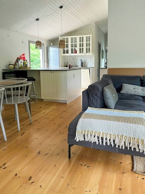 A cozy living area with a sofa, a dining table with chairs, and a kitchen with white cabinets and a window. - Summer house on nature plot near Lynæs (Hundested)