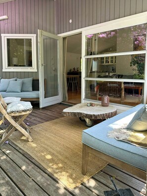 Un patio ouvert avec une terrasse en bois, un coin salon agrémenté de coussins, une table au plateau rustique et une vue sur l’intérieur de la maison grâce à une porte vitrée.