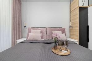 Ein modernes Schlafzimmer mit Bett, Kissen, Nachttisch, Wand-Fernseher und einem Fenster mit Vorhängen.