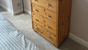 A wooden chest of drawers in a room with a carpeted floor and a window with blinds. - London Charm - 3 Bedroom House (London)