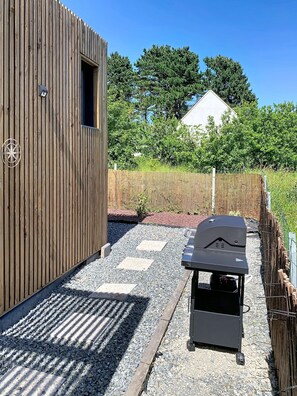 Una construcción de madera con una ventana, una parrilla para barbacoa y un camino de grava.