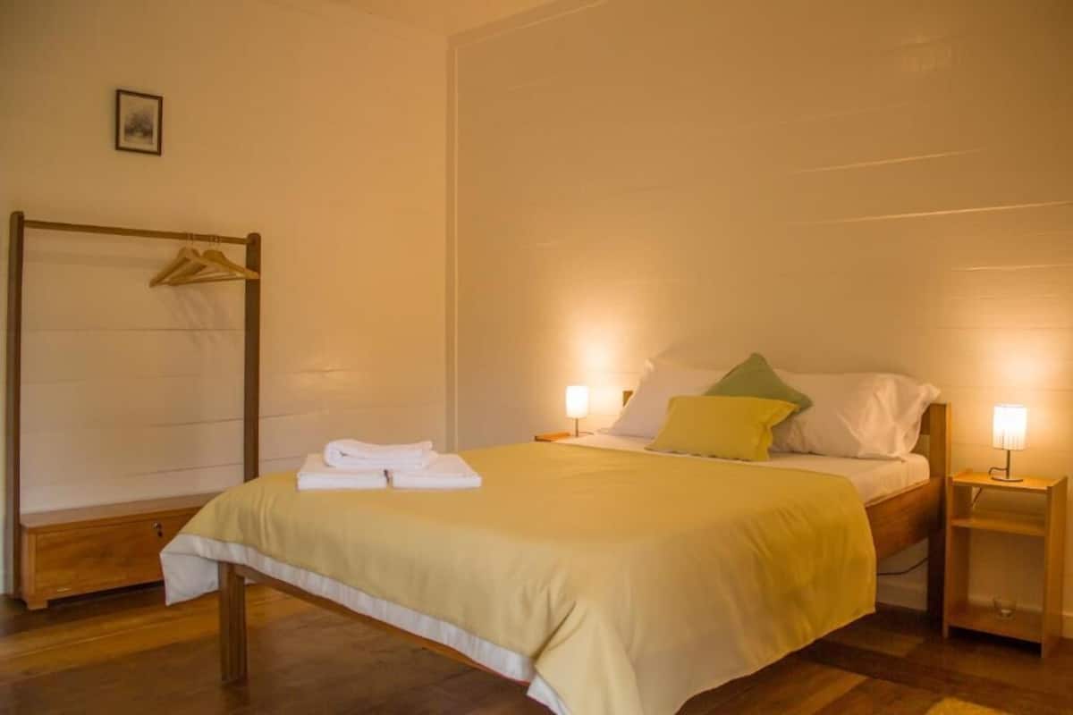 A single bed with a yellow bedspread, two bedside lamps, a wooden nightstand, and a wooden bench with a towel.