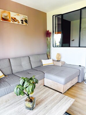 Ein modernes Wohnzimmer mit einem Ecksofa, einem Couchtisch, einer Zimmerpflanze und einem großen Fenster.