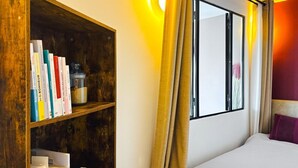 Ein Hotelzimmer mit einem Holzbücherregal, einem Bett, einem Fenster mit Vorhängen und einer Wandlampe.