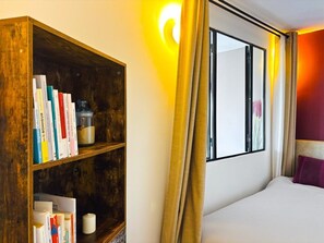 Ein Hotelzimmer mit einem Holzbücherregal, einem Bett, einem Fenster mit Vorhängen und einer Wandlampe.