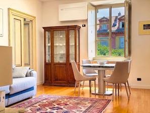 Ein Zimmer mit Esstisch, Stühlen, einer Couch, einer Vitrine und einem Fenster mit Blick auf Gebäude.