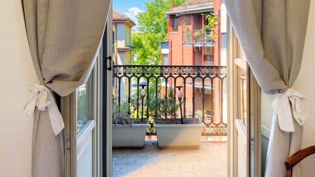 Uitzicht door een raam met teruggebonden gordijnen, waarop een balkon met een balustrade, potplanten en een gebouw met balkons te zien is.
