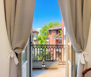 A view through a window with curtains tied back, revealing a balcony with a railing, potted plants, and a building with balconies in the background. - Casa Paravia - History meets contemporary comfort (Torino)