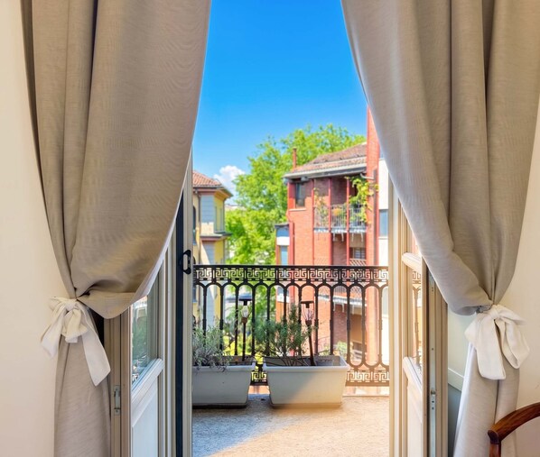 Blick durch ein Fenster mit zurückgebundenen Vorhängen auf einen Balkon mit Geländer, Pflanzgefäßen und ein Gebäude mit Balkonen im Hintergrund.