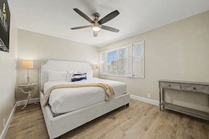 Un dormitorio con cama, ventilador de techo, mesita de noche con lámpara, ventana con persianas y cómoda.