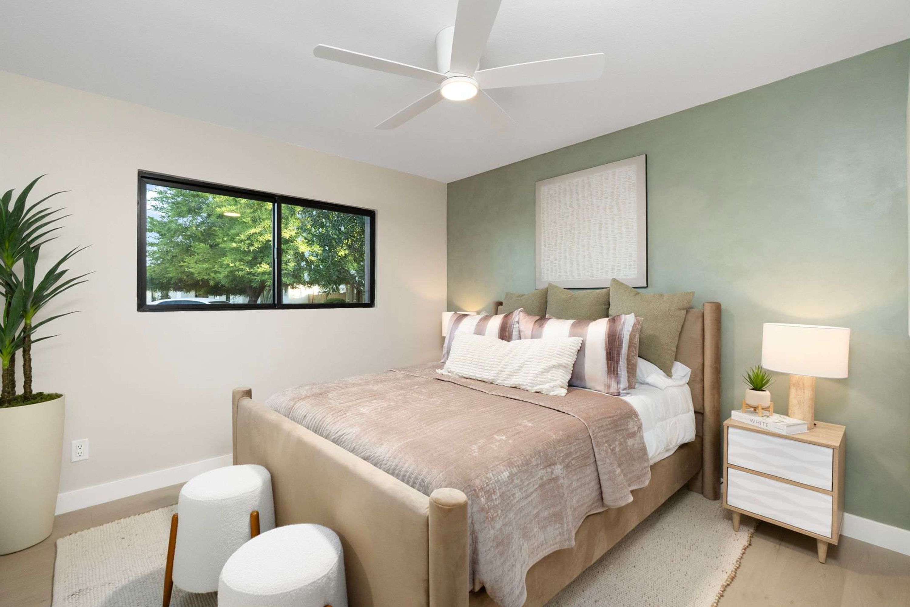 Une chambre à coucher comprenant un lit, une table de chevet, une commode, un ventilateur de plafond, une fenêtre et une plante en pot.