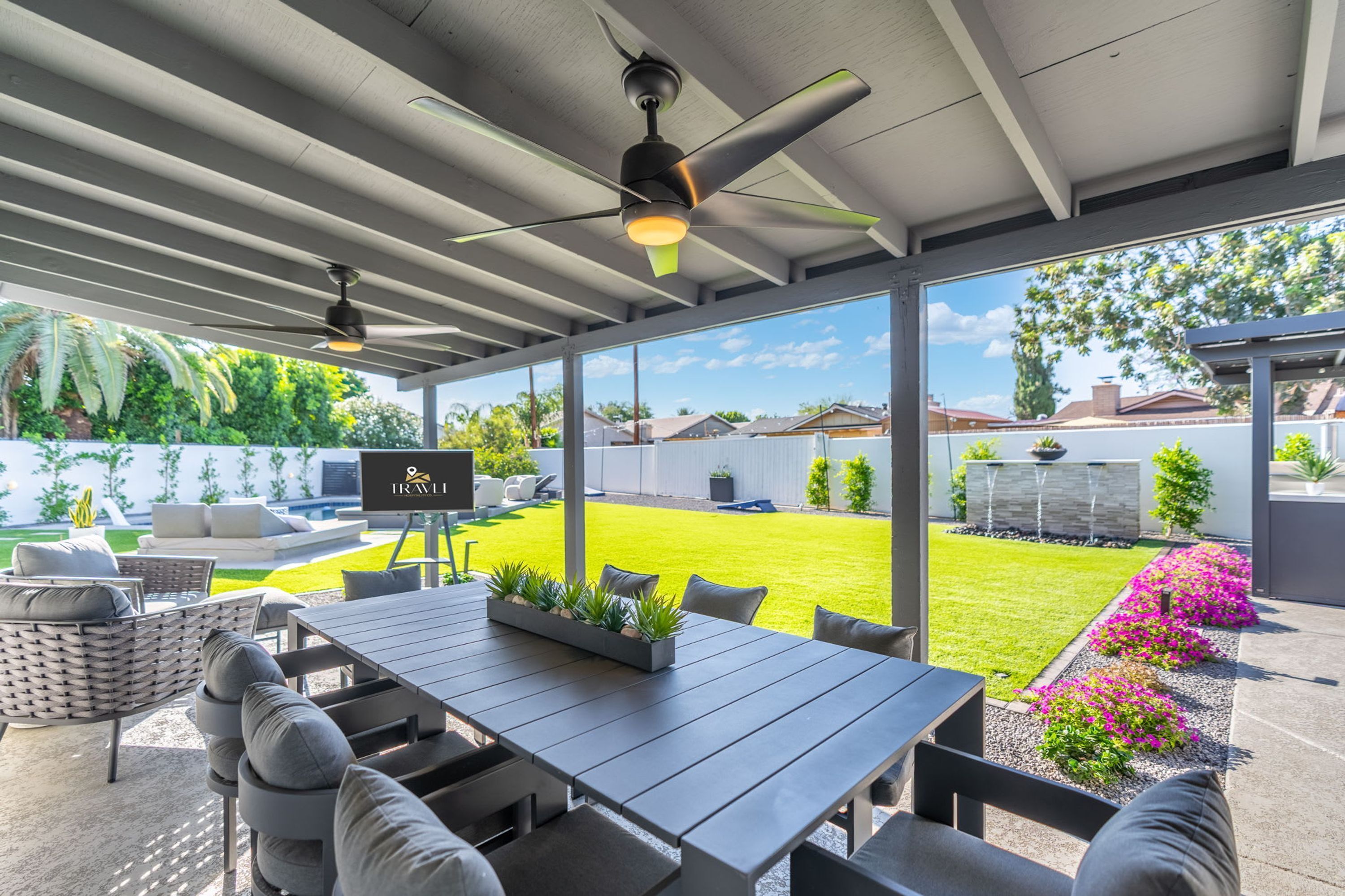 An outdoor patio with a table and chairs, a ceiling fan, and a view of a well-maintained lawn and garden.
