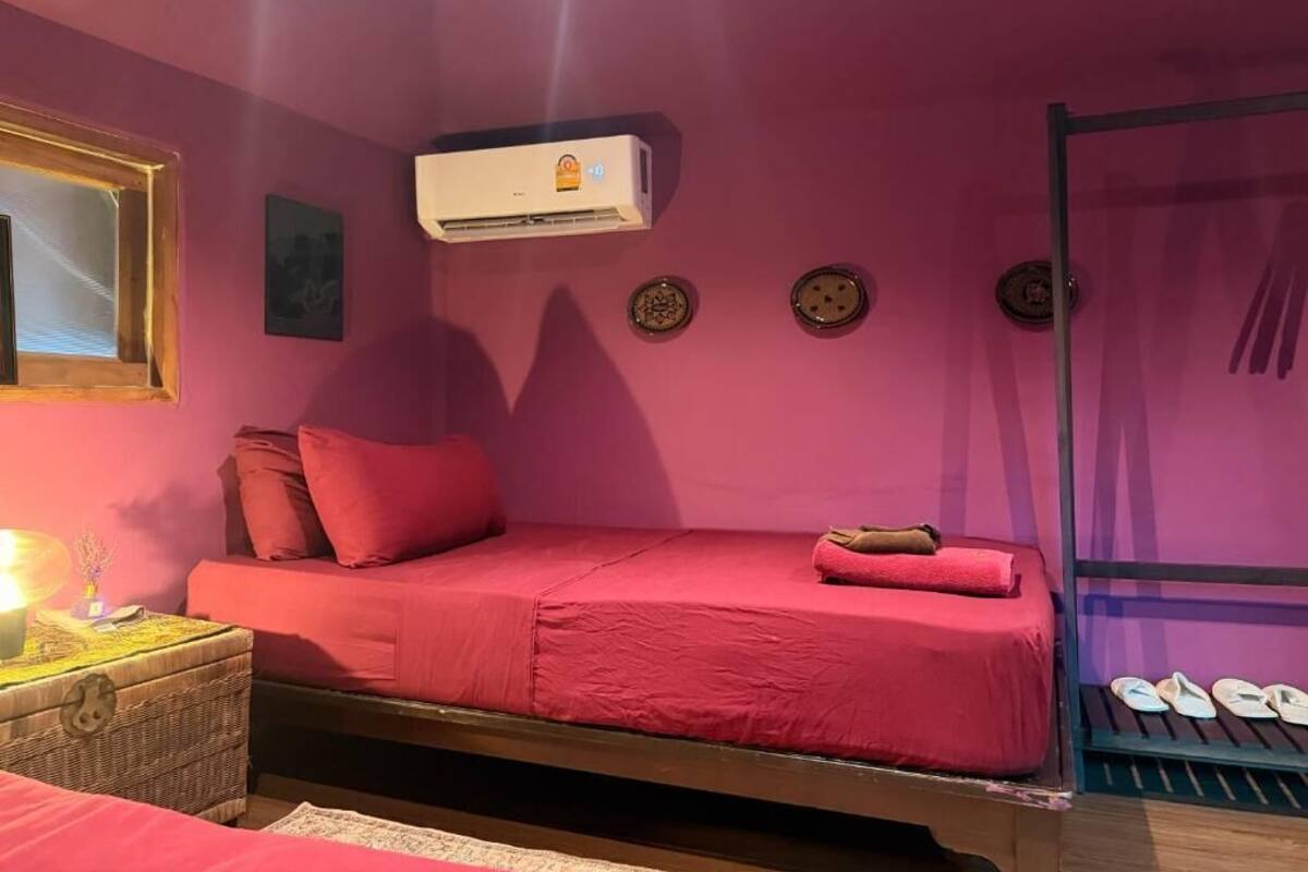 A room with a bed, a small table with a lamp, an air conditioner, and a shoe rack.