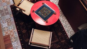 Ein Zimmer mit einem gemusterten Boden, mehreren Sitzbereichen inklusive Stühle und einem Sofa, einem roten Regenschirm und einem kleinen Tisch mit einer Zeitschrift.